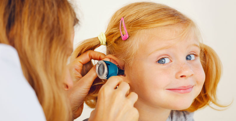 Ein kleines Mädchen schaut lächeln in die Kamera, während ihr Ohr untersucht wird.